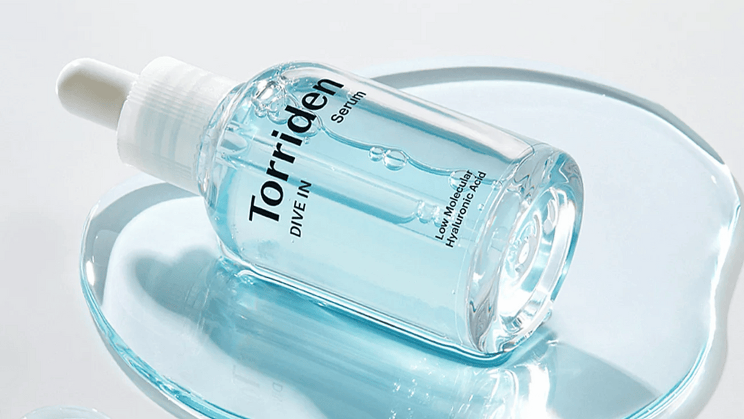 En genomskinlig droppflaska med Torriden Dive In Serum, fylld med en ljusblå hyaluronsyra-lösning, placerad på en matchande blå glasskiva som reflekterar ljuset.