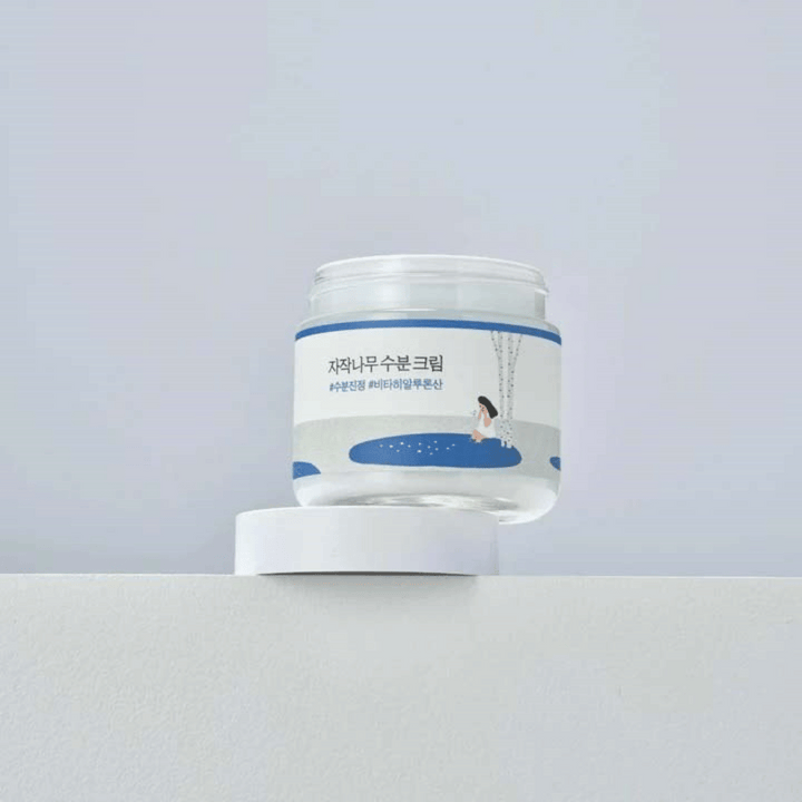 Round Lab Birch Juice Moisturizing Cream i transparent burk med vit etikett och blå illustration av kvinna vid björk, stående på vit yta mot ljus bakgrund.