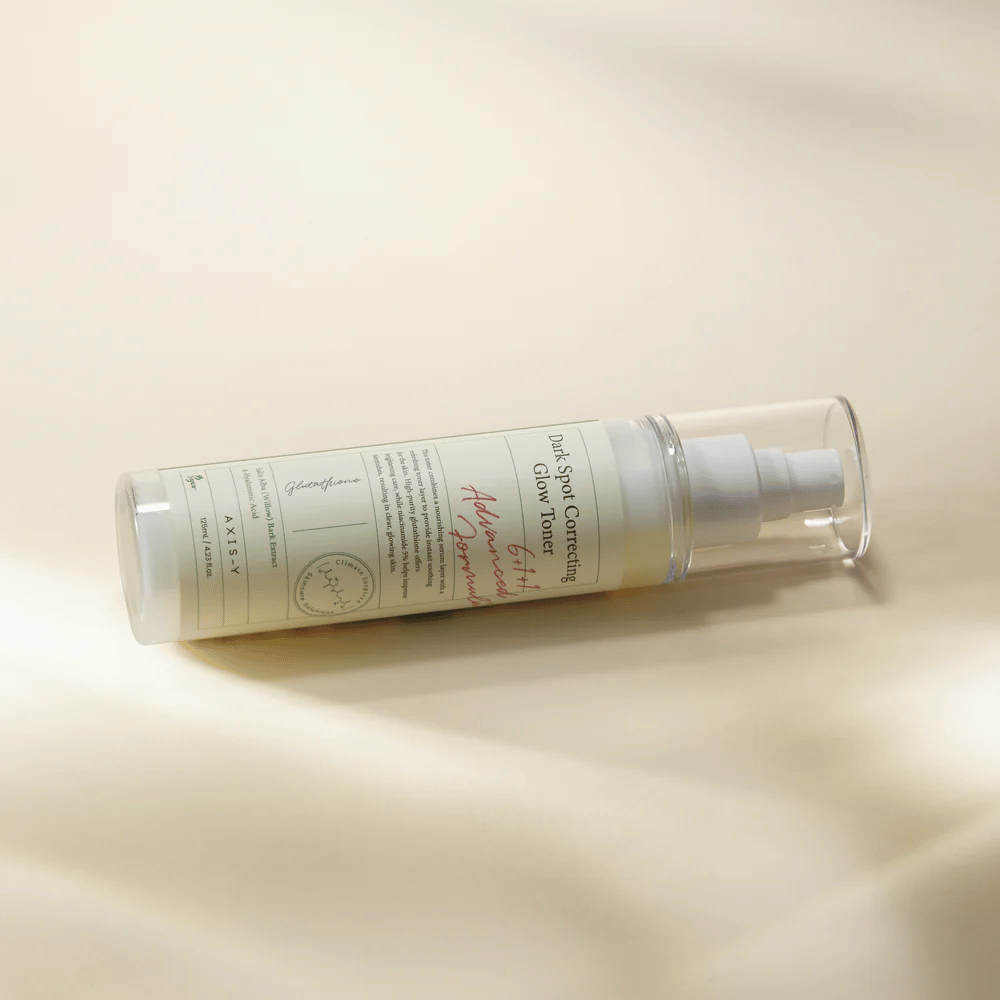 En liggande flaska av "AXIS-Y Dark Spot Correcting Glow Toner" placerad på en ljus yta. Flaskan är genomskinlig med en vit etikett och har en vit spraypump. Etiketten visar information om produktens ljusnande och återfuktande egenskaper samt innehåller nyckelingredienser som Glutathione och Niacinamide.