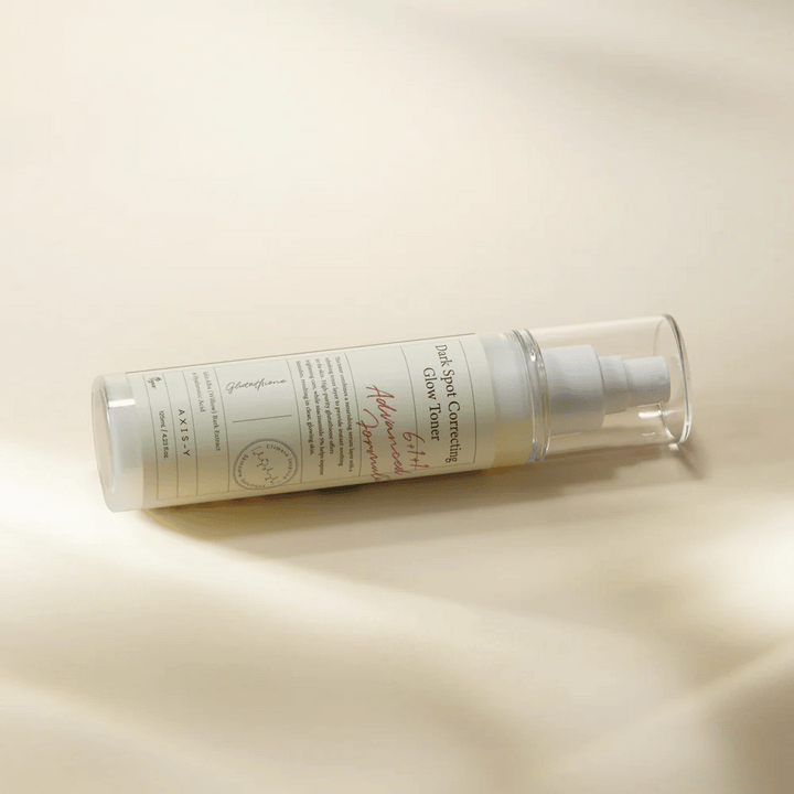 En liggande flaska av "AXIS-Y Dark Spot Correcting Glow Toner" placerad på en ljus yta. Flaskan är genomskinlig med en vit etikett och har en vit spraypump. Etiketten visar information om produktens ljusnande och återfuktande egenskaper samt innehåller nyckelingredienser som Glutathione och Niacinamide.