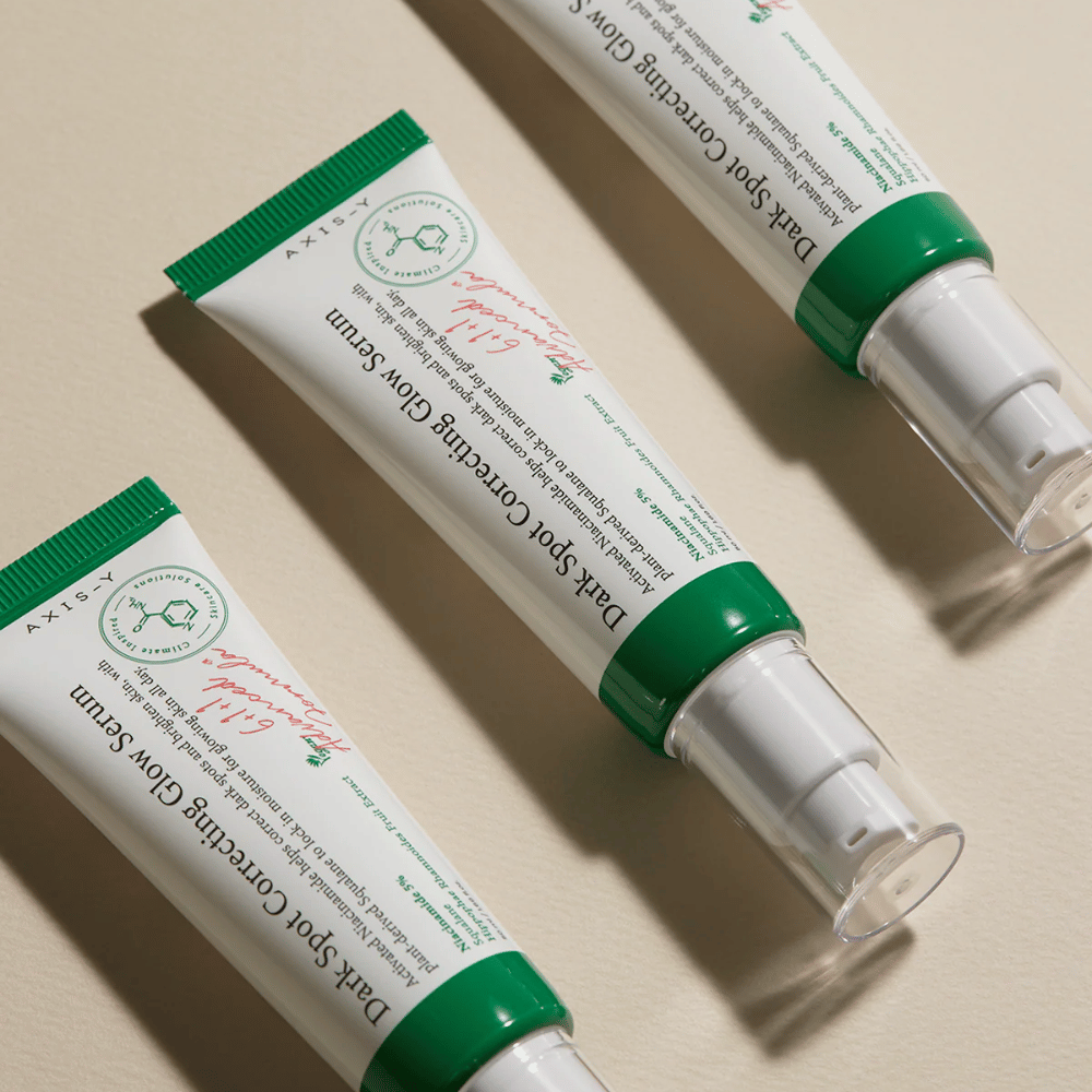 Tre tuber av Axis-Y Dark Spot Correcting Glow Serum med grön kork och vit förpackning ligger på ett ljust underlag.