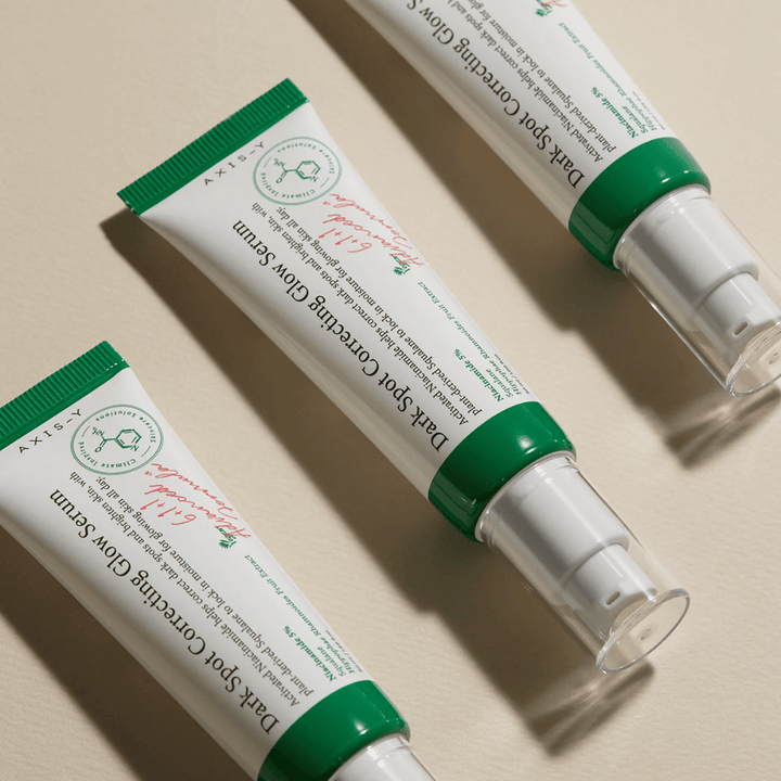 Tre tuber av Axis-Y Dark Spot Correcting Glow Serum med grön kork och vit förpackning ligger på ett ljust underlag.