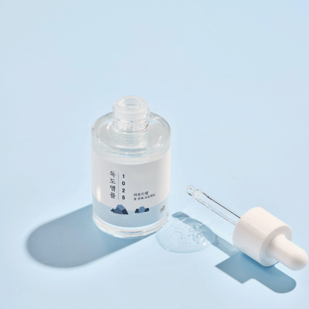 Una botella abierta de 1025 Dokdo Ampoule con un gotero al lado, contra un fondo azul que crea una sombra suave.