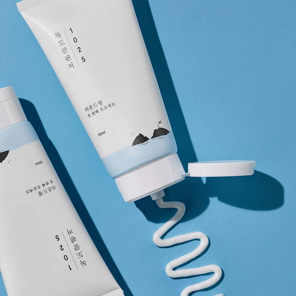 Dos tubos de 1025 Dokdo Cleanser sobre fondo azul, uno con tapa abierta y una línea ondulada de limpieza exprimida.