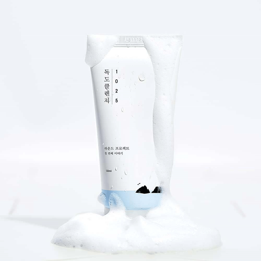 1025 Dokdo Cleanser en un tubo blanco cubierto de espuma, con gotas de agua y espuma alrededor de la base, aislado sobre fondo claro.