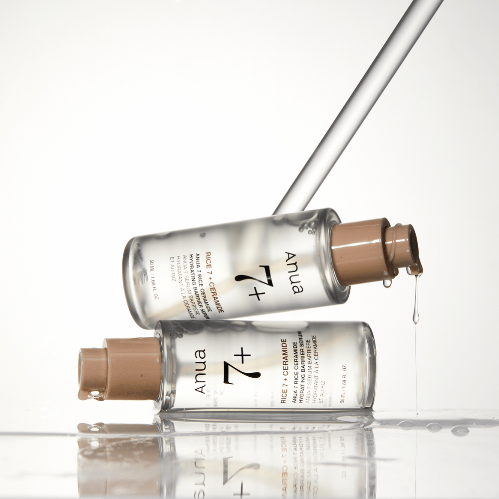 La imagen muestra dos botellas transparentes de "Anua 77 Rice + Ceramide Hydrating Barrier Serum" con gotas que caen de una pipeta. El suero, que está enriquecido con arroz y ceramidas, está destinado a hidratar y fortalecer la barrera de la piel. Las botellas están elegantemente diseñadas, lo que refuerza la sensación premium del producto.
