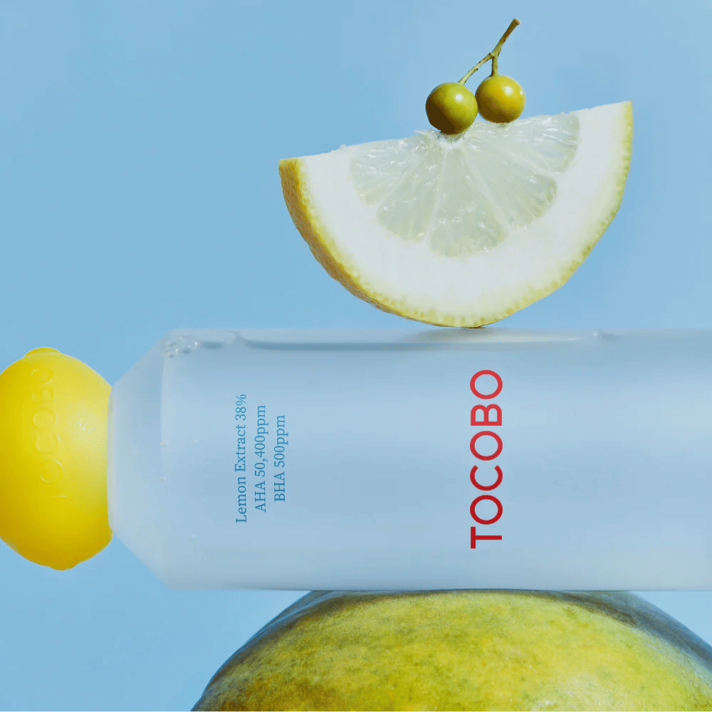 Botella con TOCOBO AHA BHA Lemon Toner, decorada con una tapa en forma de limón. Una rodaja de limón y pequeñas bayas verdes equilibran sobre la botella contra un fondo azul.