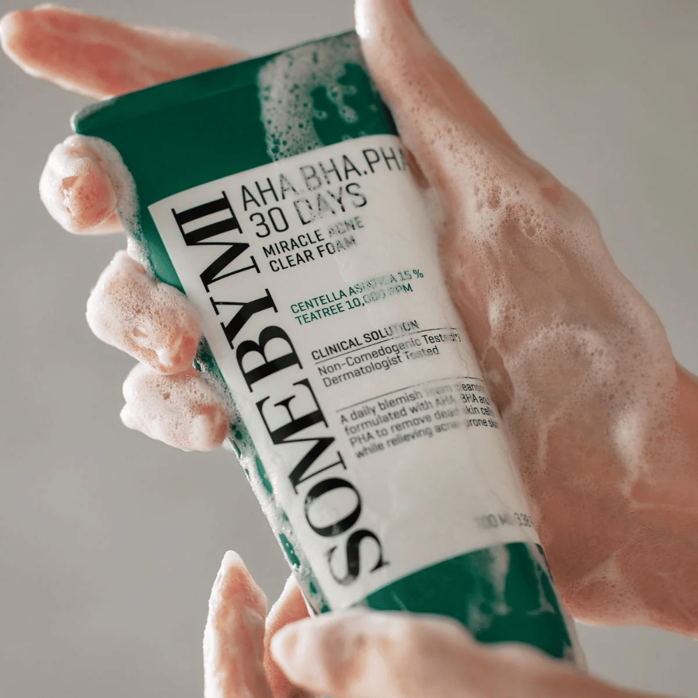 Una persona sostiene un tubo de "SOME BY MI AHA, BHA, PHA 30 Days Miracle Acne Clear Foam" con contenido espumoso sobre las manos. El producto está destinado para uso diario para combatir el acné y limpiar los poros con sus ingredientes activos como centella, aceite de tea tree y ácidos como AHA, BHA y PHA.