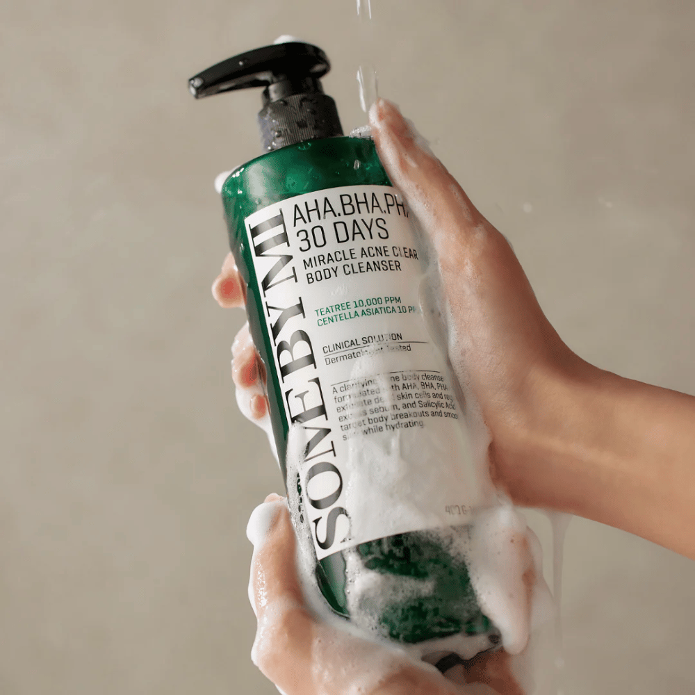 Una persona sostiene una botella SOME BY MI AHA.BHA.PHA 30 Days Miracle Acne Clear Body Cleanser. La botella es verde con una etiqueta blanca y está cubierta de espuma del limpiador. La persona se lava con el producto mientras el agua corre sobre sus manos.