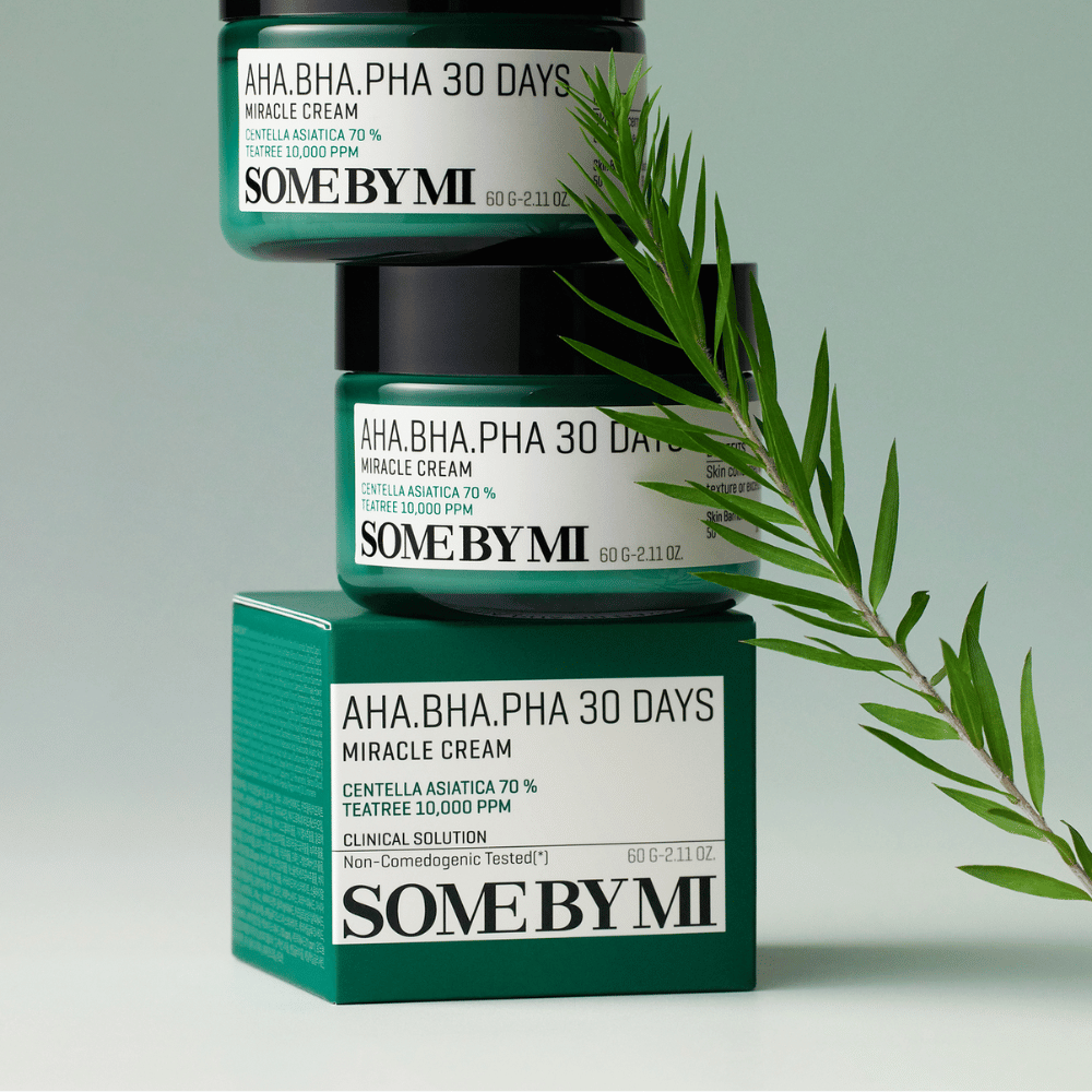 Una imagen que muestra dos frascos de "SOME BY MI AHA, BHA, PHA 30 Days Miracle Cream" apilados sobre una caja de tono verde. La crema, que es rica en centella asiática y aceite de tea tree, está diseñada para mejorar la calidad de la piel y aliviar problemas cutáneos. Actúa hidratando y calmando.