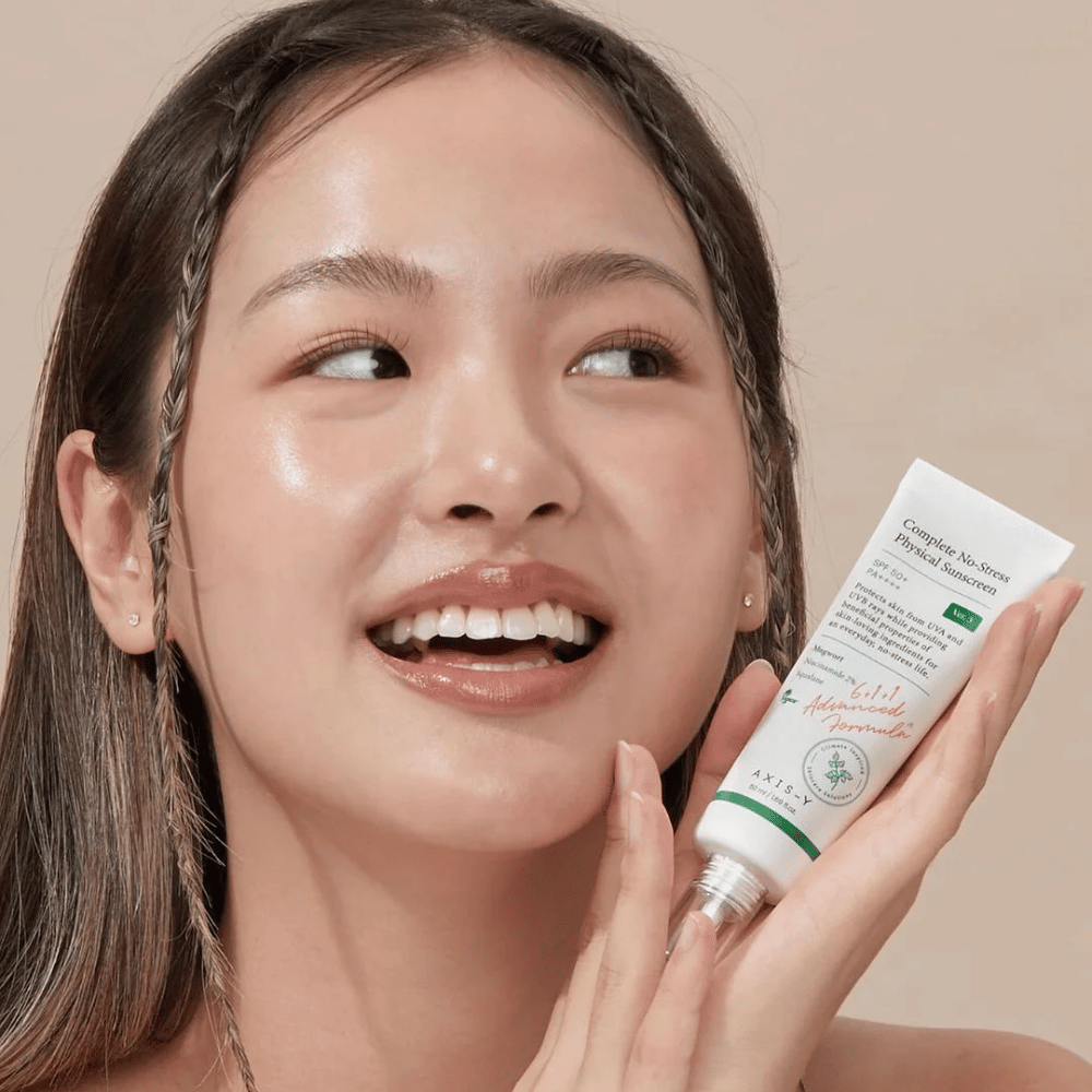 La imagen muestra a una joven sonriendo alegremente y sosteniendo un tubo con AXIS-Y Complete No-Stress Physical Sunscreen. Ella tiene un aspecto radiante y natural con un maquillaje mínimo, lo que resalta el producto que presenta.