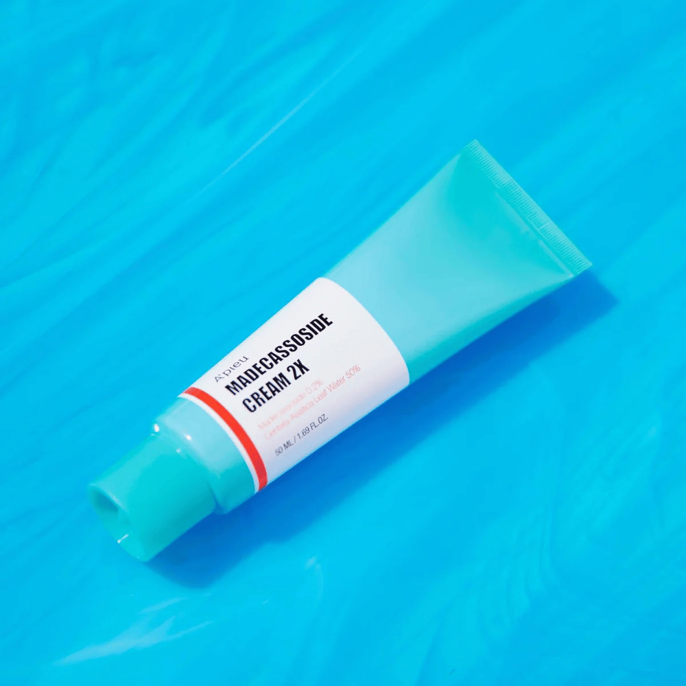 Un tubo A'PIEU Madecassoside Cream 2X está colocado en diagonal sobre un fondo azul con arrugas similares al agua. El tubo tiene un cuerpo blanco con una tapa turquesa clara y una etiqueta de color rojo a naranja donde el nombre del producto y el ingrediente activo Madecassoside 2% están claramente impresos. En la etiqueta también se indica la cantidad que es 50 ml / 1.8 fl oz. La presentación brillante y colorida da una sensación fresca y energética.