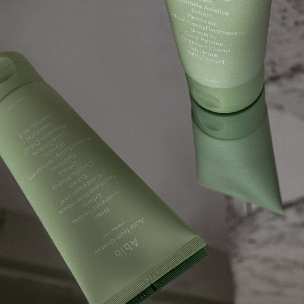 Tubo verde de Abib Acne Foam Cleanser Heartleaf acostado sobre una mesa de espejo con el frente del producto y el texto reflejado. Ingredientes como ácido salicílico, centella y extracto de hoja de corazón se ven claramente en el empaque.
