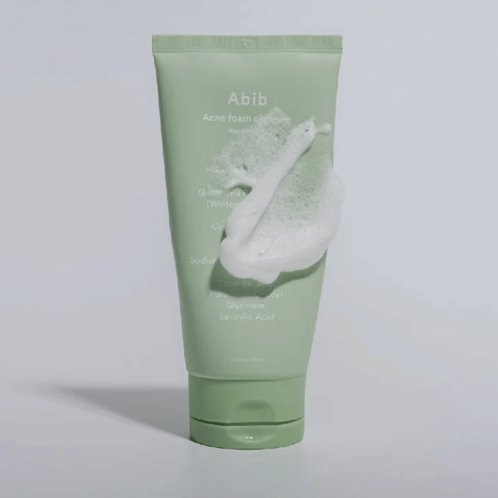 Tubo verde con Abib Acne Foam Cleanser Heartleaf, cubierto de una espuma blanca que ilustra el efecto de limpieza espumante. El producto contiene extracto de hoja de corazón y ácido salicílico para piel propensa al acné.