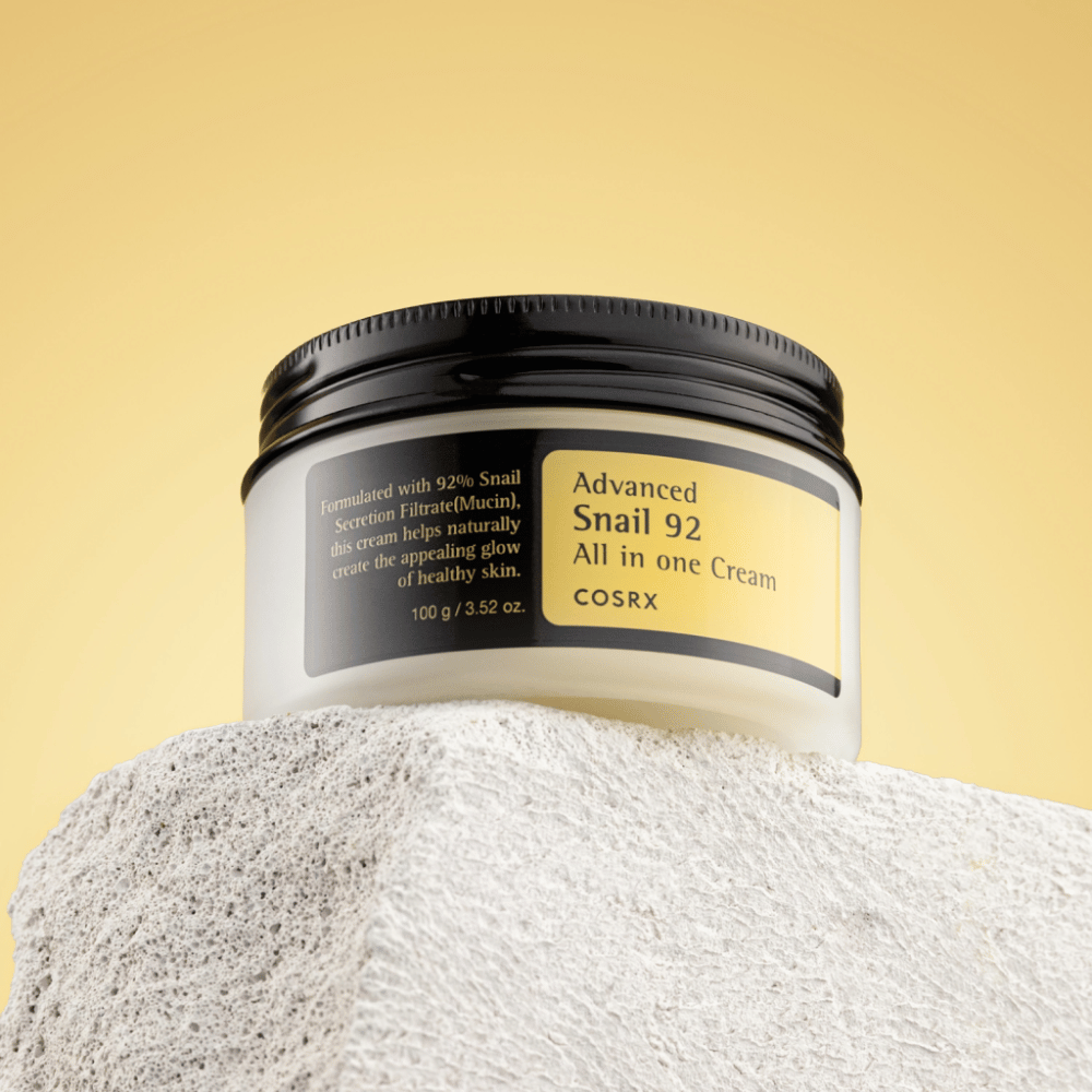 Un tarro de COSRX Advanced Snail 92 All in one Cream colocado sobre una superficie rocosa con un fondo amarillo.