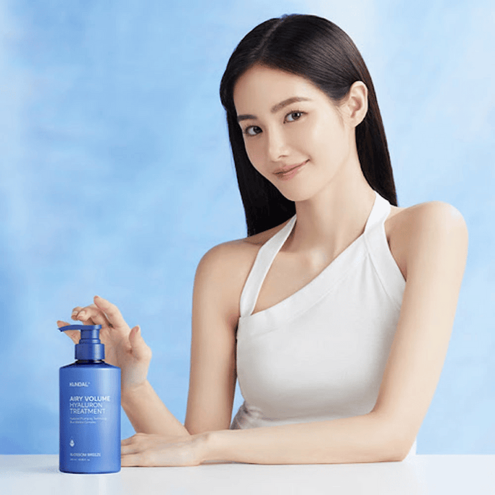 Kvinna med långt, glansigt hår poserar med KUNDAL Airy Volume Hyaluron Treatment Blossom Breeze i blå pumpflaska.