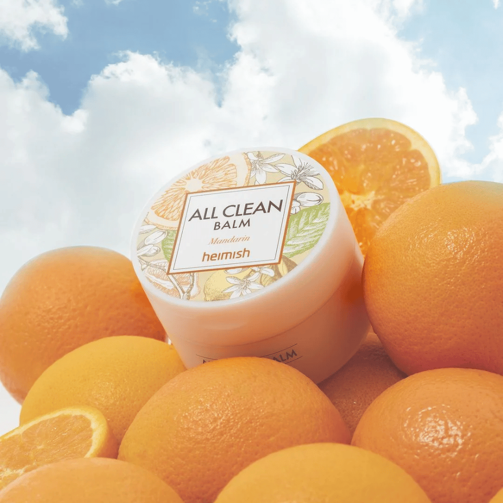 Frasco de Heimish All Clean Balm Mandarin colocado sobre una pila de naranjas, con un cielo azul de fondo.