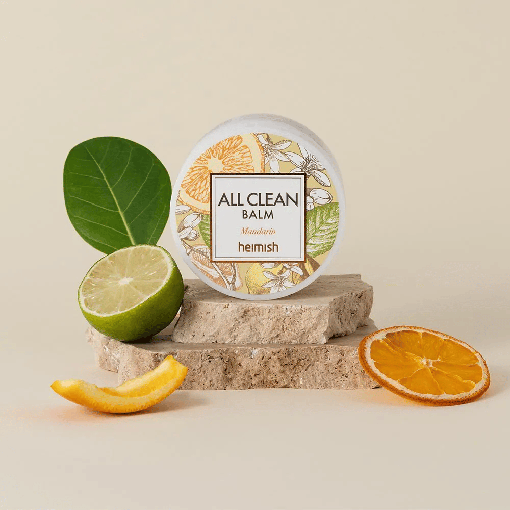 Frasco de Heimish All Clean Balm Mandarin colocado sobre piedras, rodeado de cáscaras de mandarina y hojas de cítricos, frente a un fondo claro.