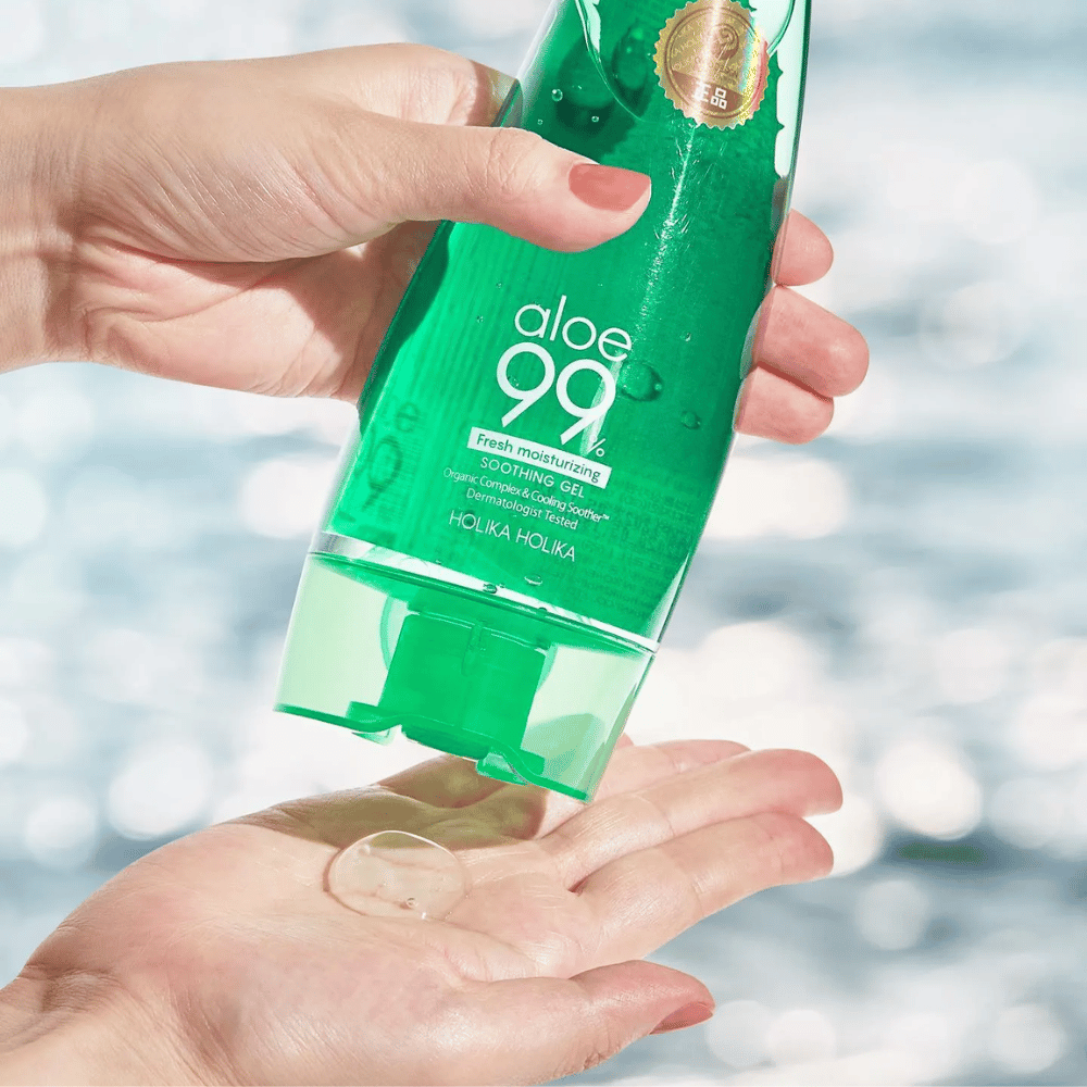 Una mano sostiene un tubo verde con Holika Holika Aloe 99% Soothing Gel, mientras otra mano captura una gota de gel que se dispensa. El fondo está borroso y muestra un patrón acuoso y brillante.