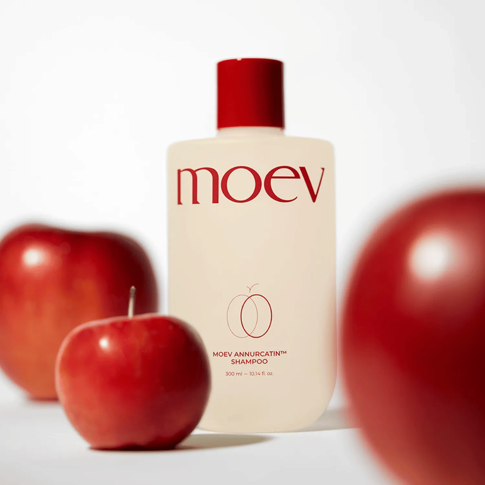 MOEV Annurcatin Shampoo colocado entre manzanas rojas – inspirado en los antioxidantes de la manzana Annurca que proporcionan un cabello más fuerte, saludable y brillante.
