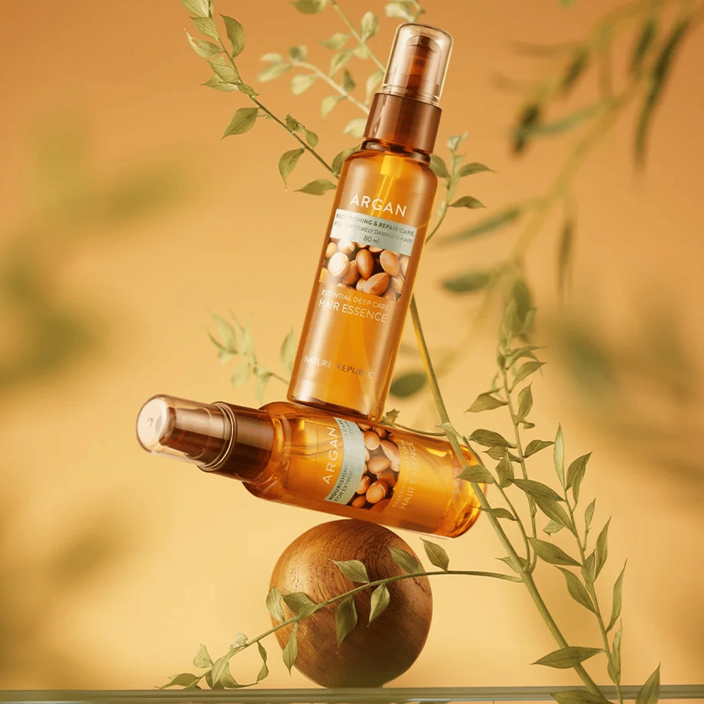 Dos botellas Nature Republic Argan Essential Moisture Hair Oil colocadas entre hojas verdes – brillo sedoso para cabello seco.