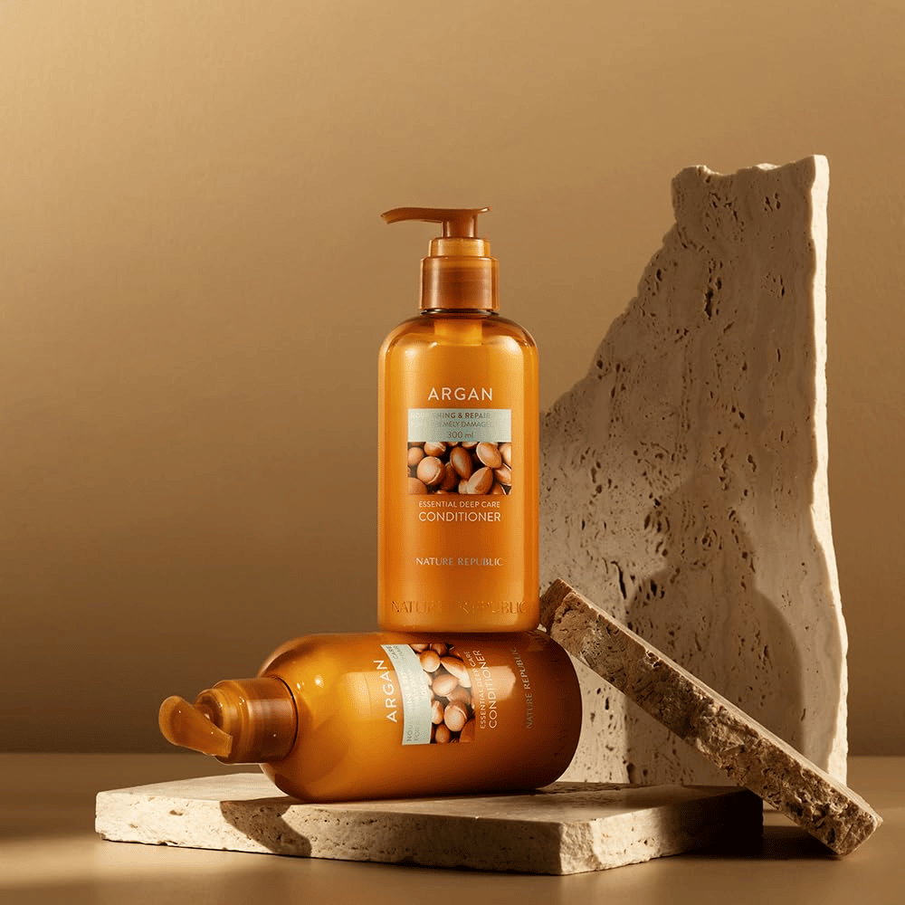 Dos botellas de Nature Republic Argan Essential Moisture Repairing Conditioner sobre una superficie de piedra en tono cálido.