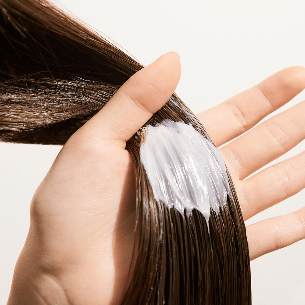 La mano aplica Nature Republic Argan Essential Moisture Repairing Hair Mask en cabello mojado – mascarilla hidratante que fortalece y suaviza el cabello.