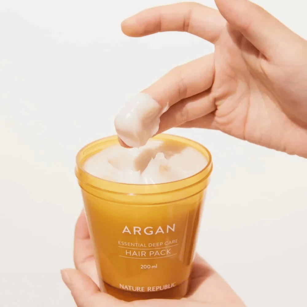 La mano toma la fórmula cremosa de Nature Republic Argan Essential Moisture Repairing Hair Mask – nutrición intensa para cabello seco.