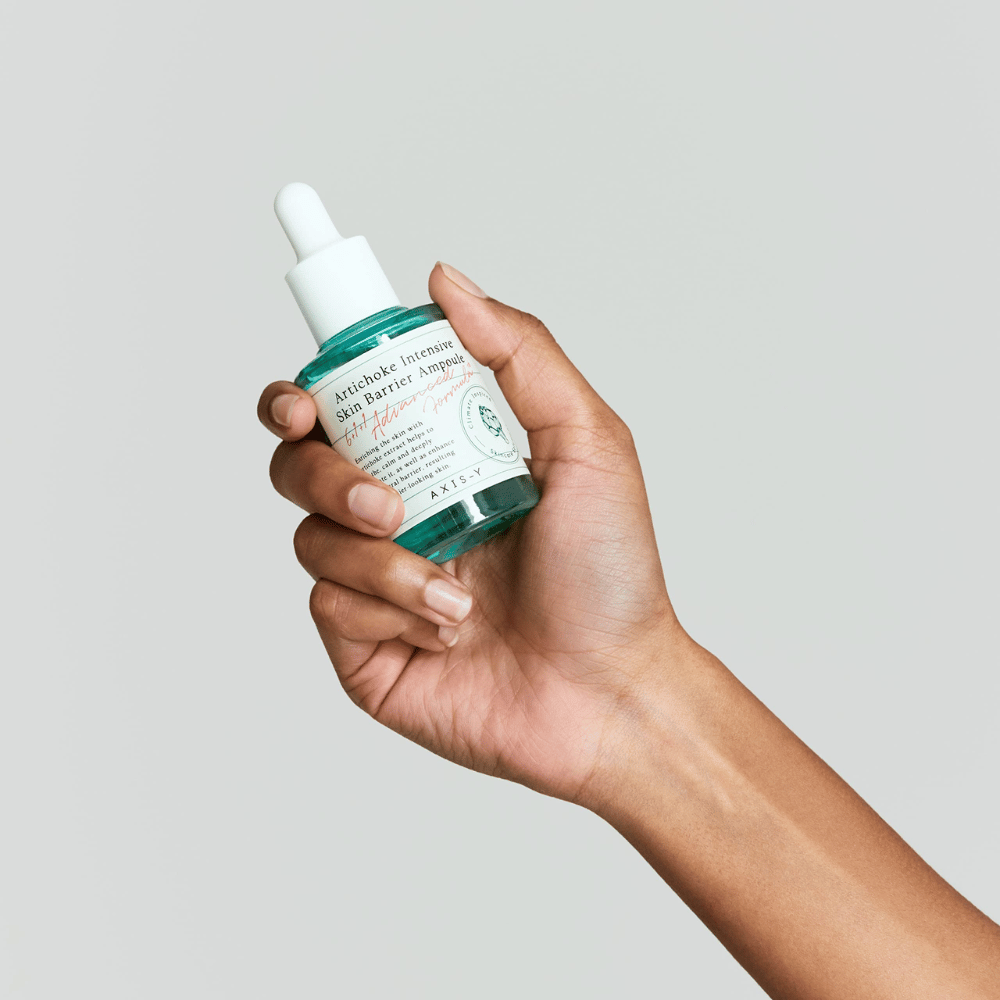 Una mano sostiene una botella de suero para el cuidado de la piel con la etiqueta "Artichoke Intensive Skin Barrier Ampoule.