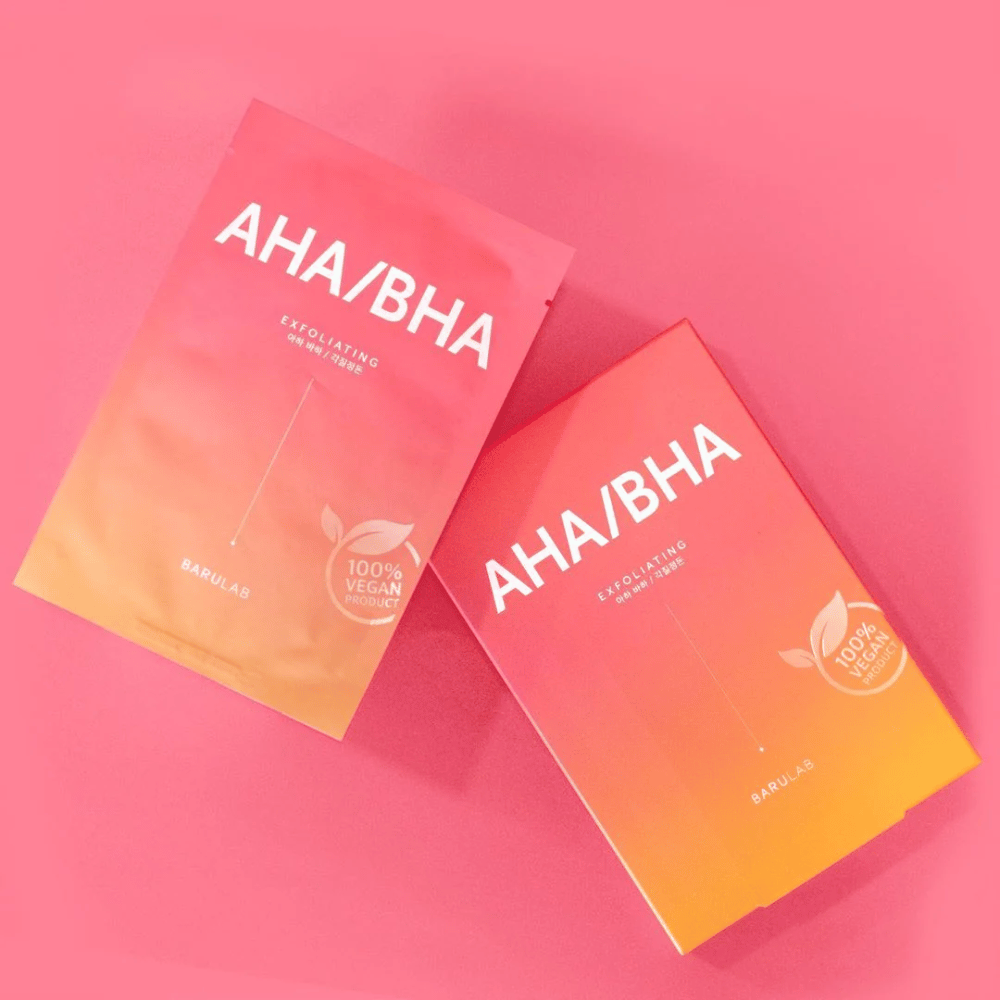 Dos productos de BARULAB, uno en una bolsa y otro en una caja, ambos marcados con "AHA/BHA" y "Exfoliante" contra un fondo rosa. Los productos están marcados como "Producto 100% Vegano".