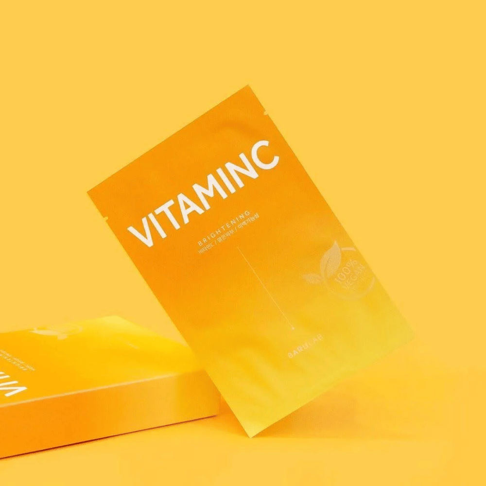 Una pila de paquetes de máscaras BARULAB "VITAMINC Brightening", todas etiquetadas como "100% Vegan Product". Los paquetes tienen un tono de color en diferentes matices de naranja y amarillo, colocados contra un fondo amarillo uniforme.