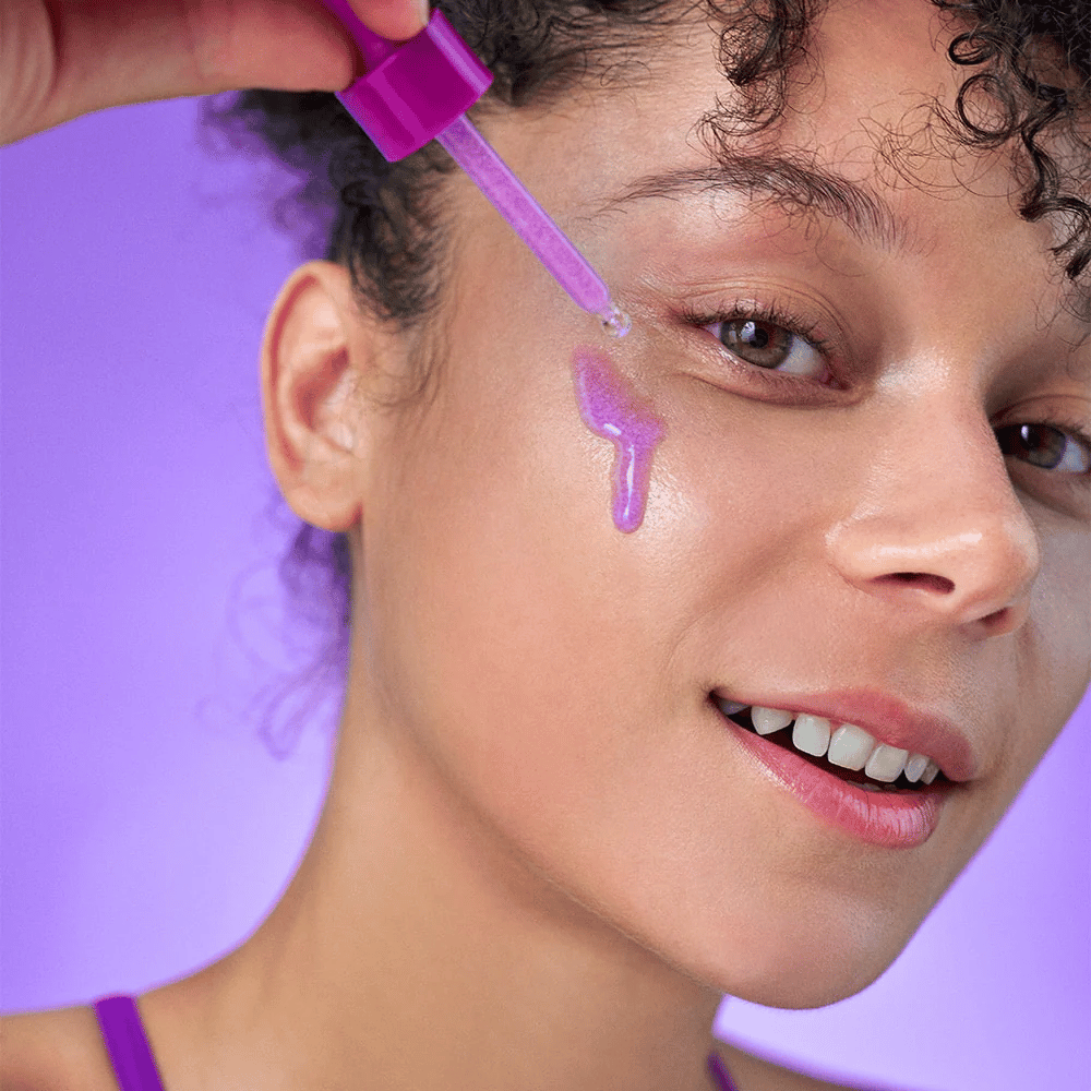 Mujer sonriente aplica un suero brillante de color lila en la piel con una pipeta. Piel radiante y fondo lila que coincide con el tono del producto.