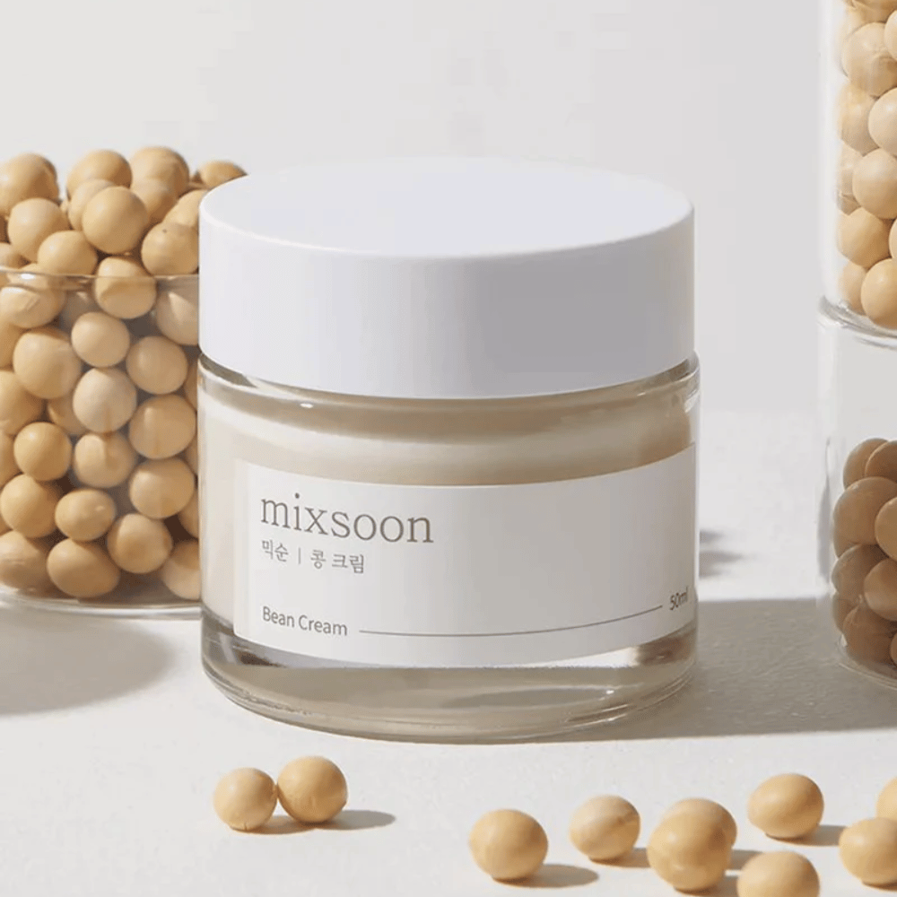 Un frasco transparente con etiqueta y tapa blanca, etiquetado "mixsoon 팥 크림" y "Bean Cream" con un volumen indicado de "50ml". El frasco contiene una crema de color amarillo claro y está colocado sobre una superficie clara. Al lado del frasco hay un contenedor de vidrio lleno de granos de color marrón claro que se han derramado sobre la superficie, lo que sugiere que la crema contiene extracto de frijol. El fondo y la iluminación crean una atmósfera cálida y natural.