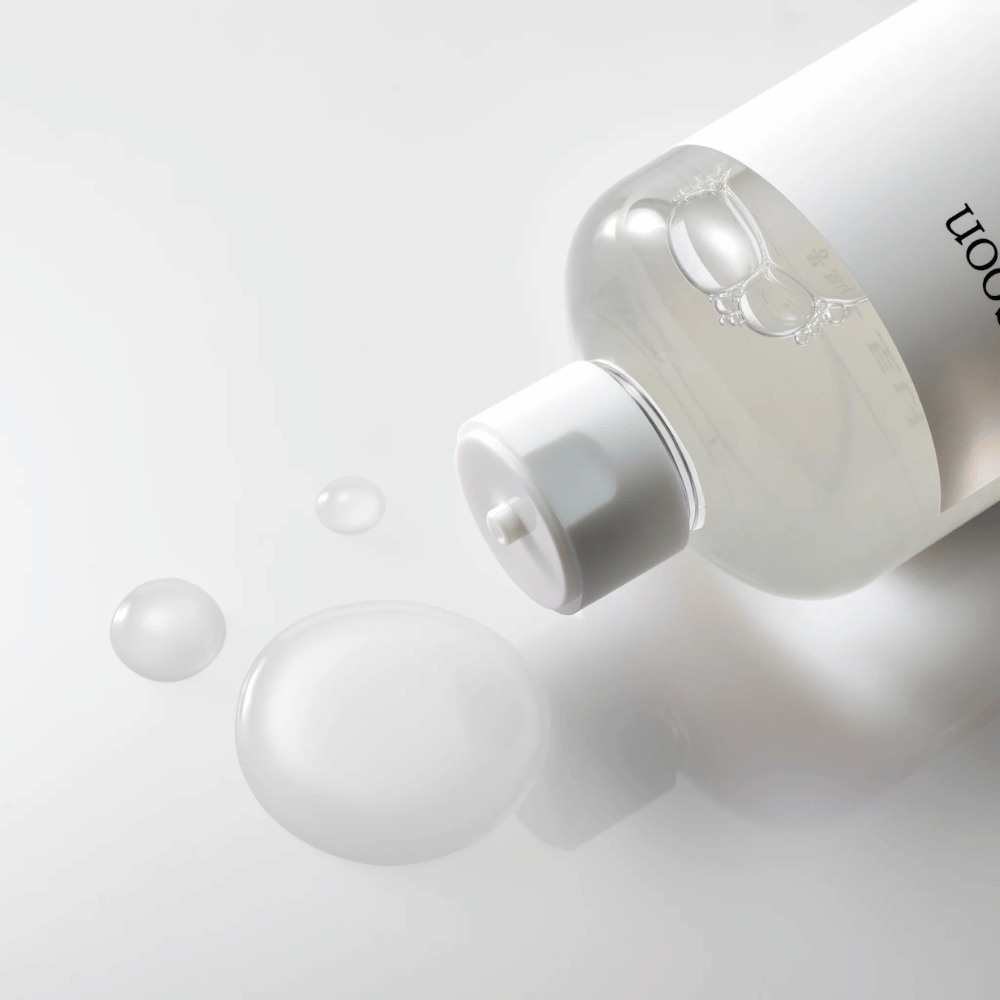 Una botella transparente yace de lado sobre una superficie brillante con un tónico ligero que lentamente gotea y forma gotas y una burbuja más grande en la superficie. La botella tiene una etiqueta blanca y un mecanismo de bomba blanca. La iluminación es suave y crea una atmósfera limpia y relajante, mientras que la consistencia fluida y claridad del tónico se destacan por cómo se esparce sobre la superficie.