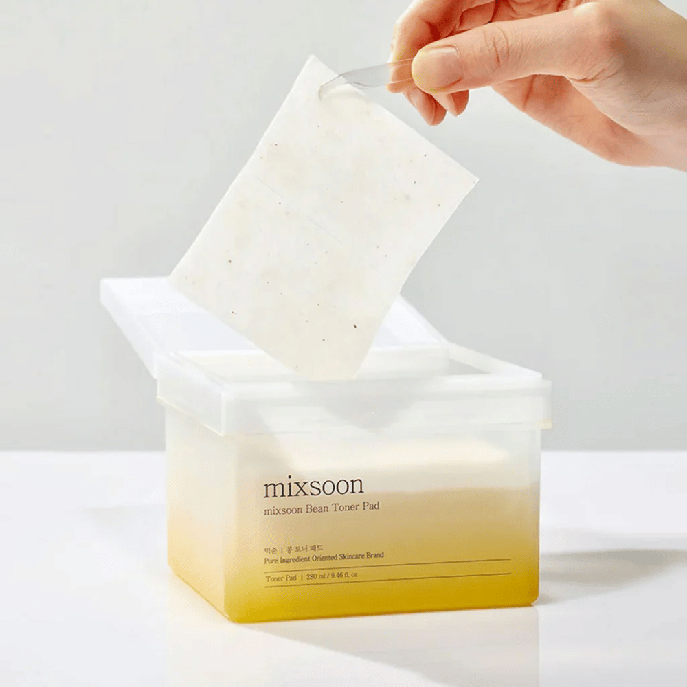 Una mano sostiene una pinza y levanta una Mixsoon Bean Toner Pad de un recipiente cuadrado y transparente con un tono amarillento. El recipiente contiene 70 almohadillas tonificantes y tiene texto que describe el producto.