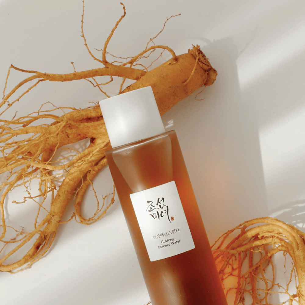 Una botella marrón Beauty of Joseon Ginseng Essence Water con tapa blanca está al lado de una raíz de ginseng fresca sobre fondo blanco.