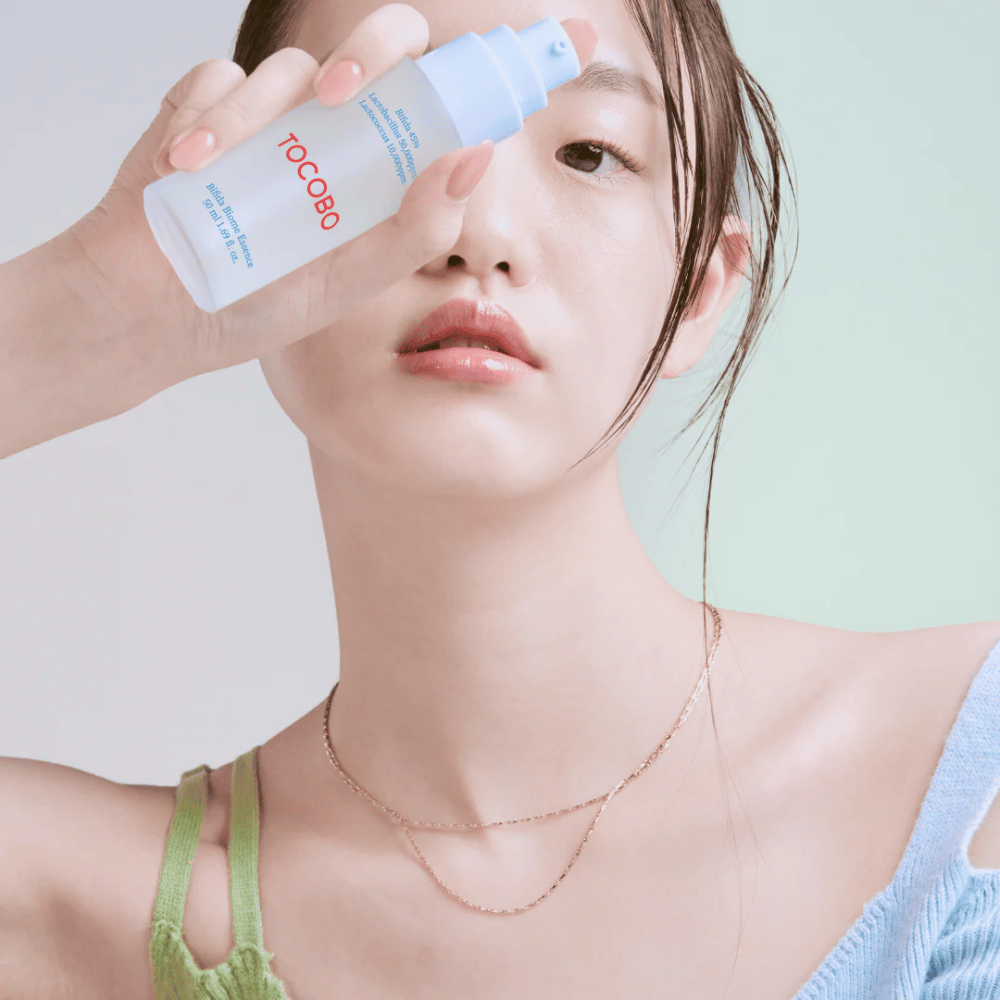 Mujer joven con maquillaje suave y tono de piel natural sostiene una botella de TOCOBO Bifida Biome Essence frente a su cara. Lleva una camiseta azul claro con tirantes verdes y un collar de oro delgado sobre un fondo claro.