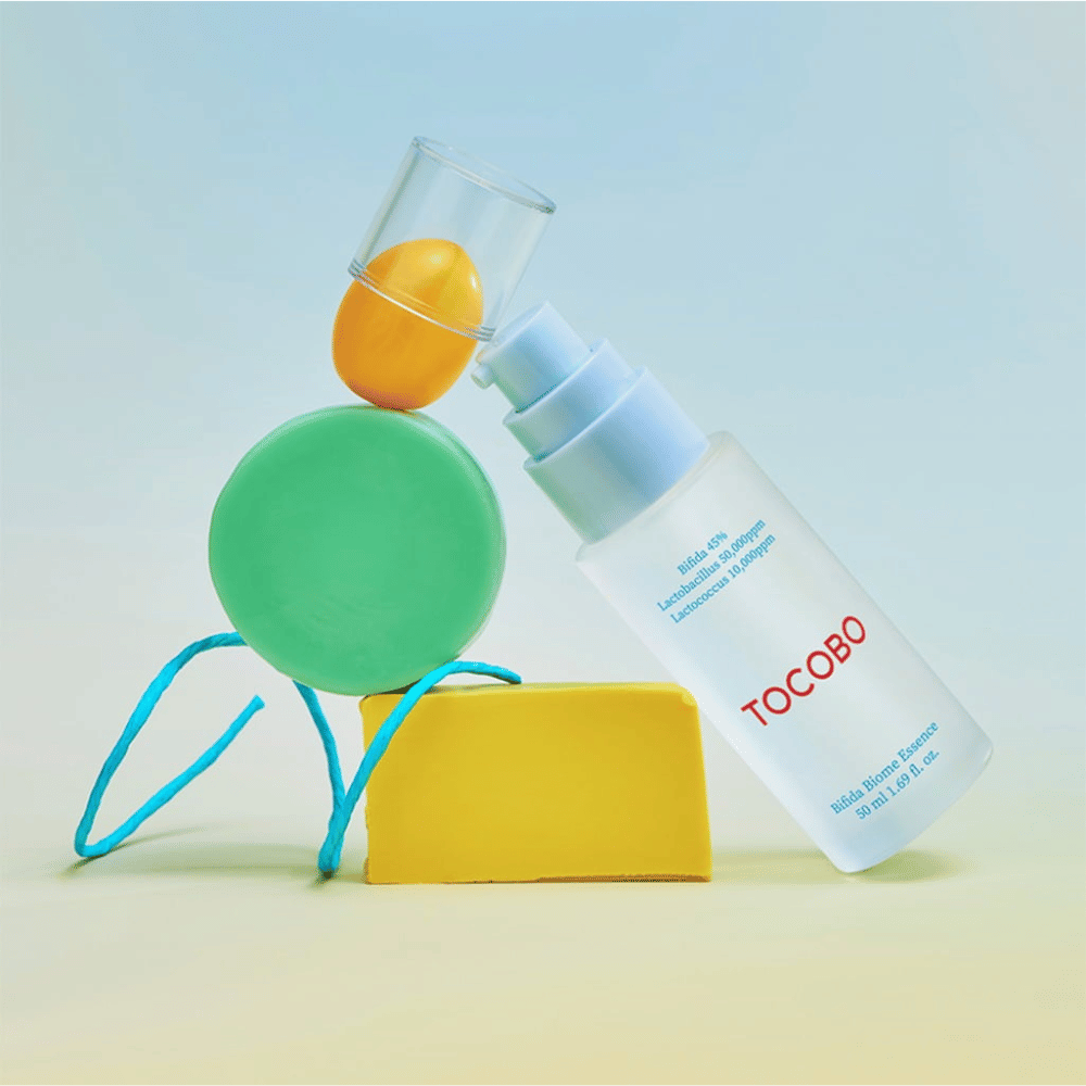 TOCOBO Bifida Biome Essence en un frasco de bomba esmerilado con logotipo rojo, colocado sobre un fondo colorido con formas geométricas verdes, amarillas y naranjas. El texto en la botella indica Bifida 5 %, Lactobacillus 3 000 ppm y Lactococcus 1 000 ppm.