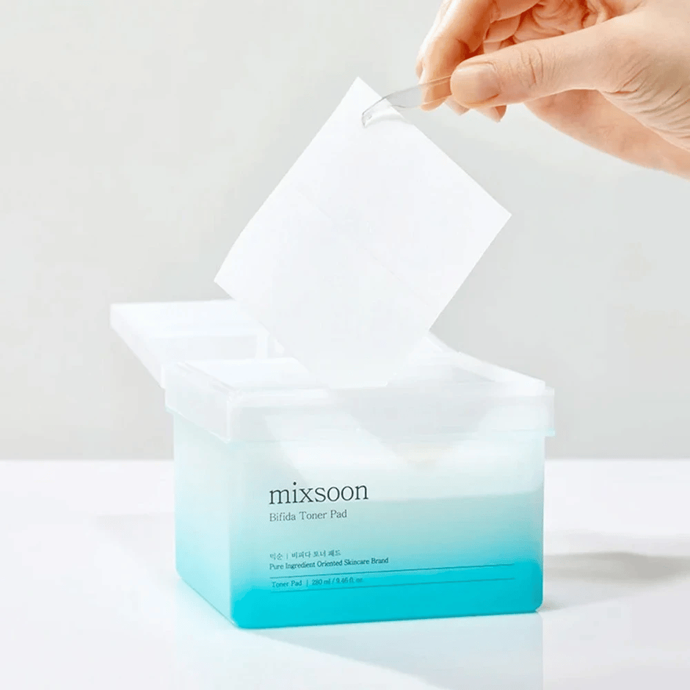 Un contenedor transparente con un degradado azul y blanco que contiene almohadillas de tónico Bifida. Una mano toma una almohadilla de tónico del contenedor, que está etiquetado con "mixsoon Bifida Toner Pad" y tiene capacidad para 280 ml (9,46 fl. oz.) de almohadillas de tónico destinadas al cuidado de la piel.