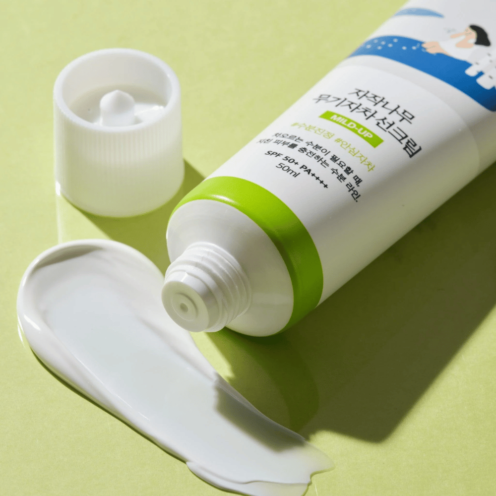 Primer plano de Birch Juice Moisturizing Mild Up Sunscreen con tapa desenroscada, crema solar blanca esparcida sobre fondo verde claro, SPF 50+ PA++++, 50 ml.