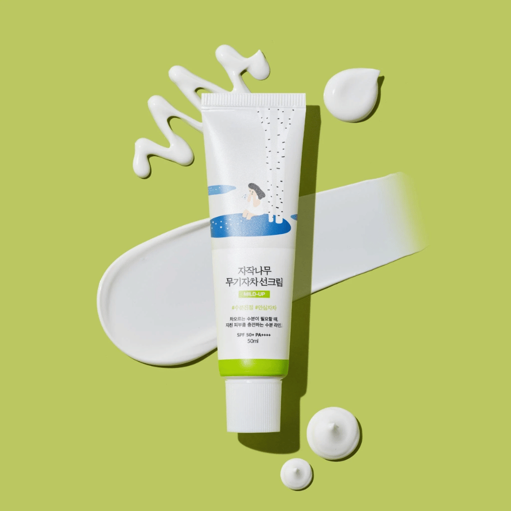 Birch Juice Moisturizing Mild Up Sunscreen – tubo blanco con base verde sobre fondo verde, rodeado de crema solar esparcida y gotas, SPF 50+ PA++++, 50 ml.