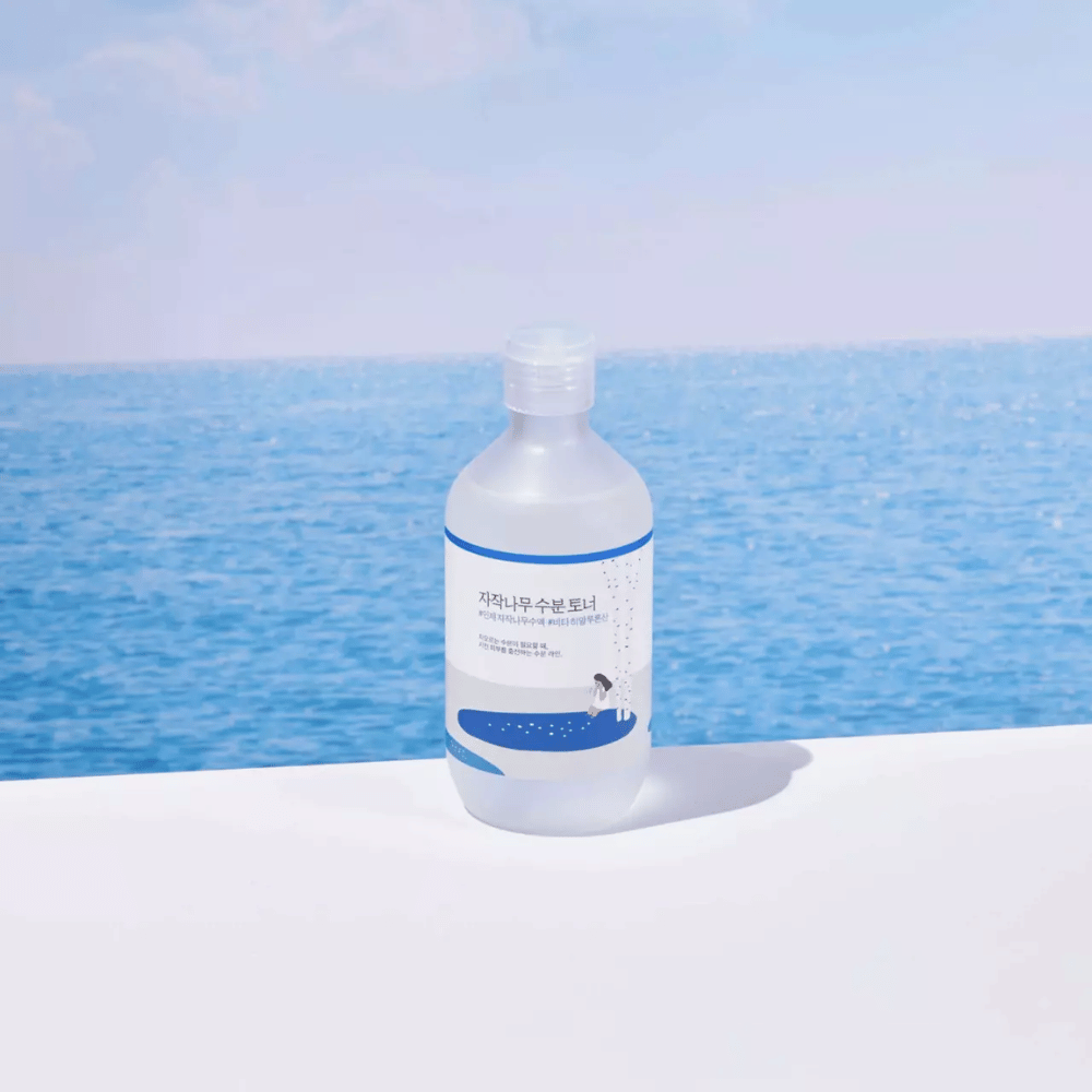 Botella de ROUND LAB Birch Juice Moisturizing Toner colocada sobre una superficie blanca con el mar y el cielo azul de fondo