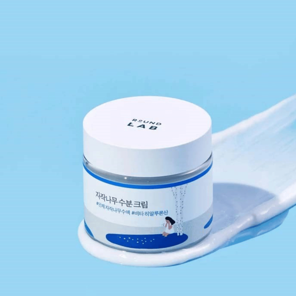 Round Lab Birch Juice Moisturizing Cream en frasco azul con tapa blanca, de pie en una capa de crema sobre fondo azul.