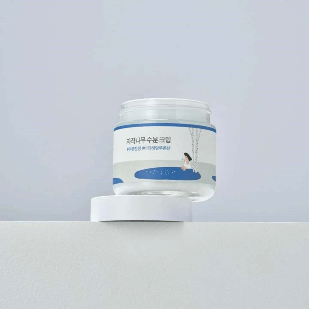 Round Lab Birch Juice Moisturizing Cream en frasco transparente con etiqueta blanca e ilustración azul de mujer junto al abedul, de pie sobre superficie blanca contra fondo claro.