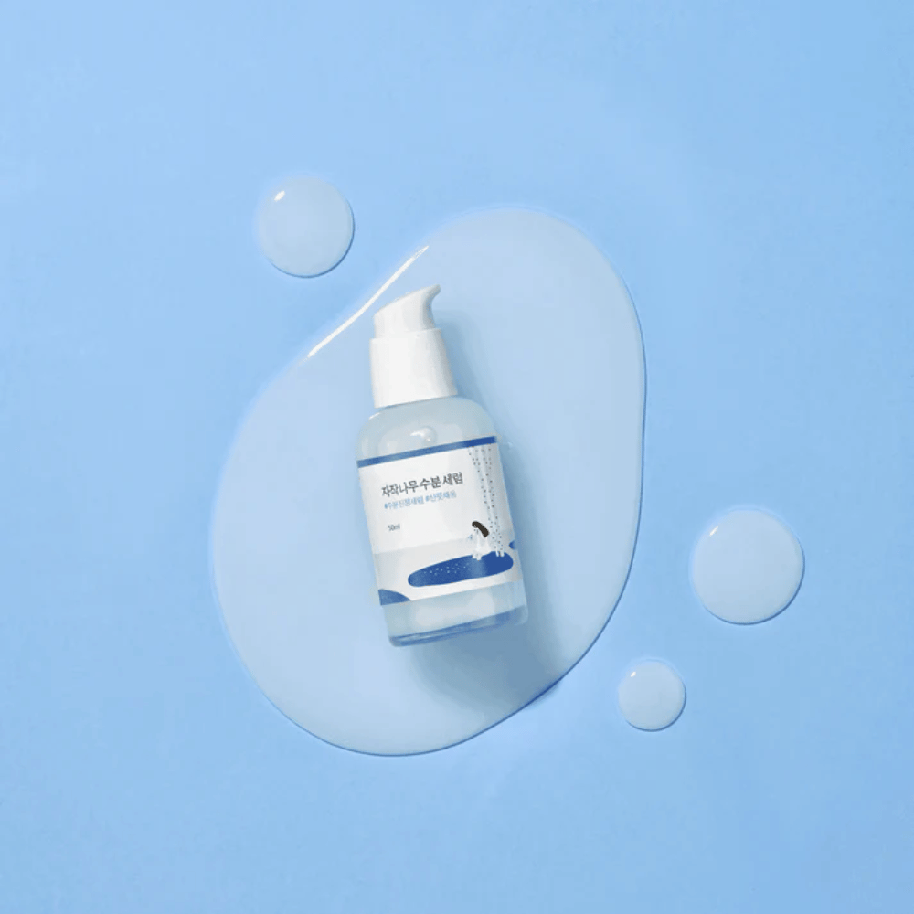 Round Lab Birch Juice Moisturizing Serum en botella de bomba transparente colocada sobre un fondo azul con gotas y suero derramado.