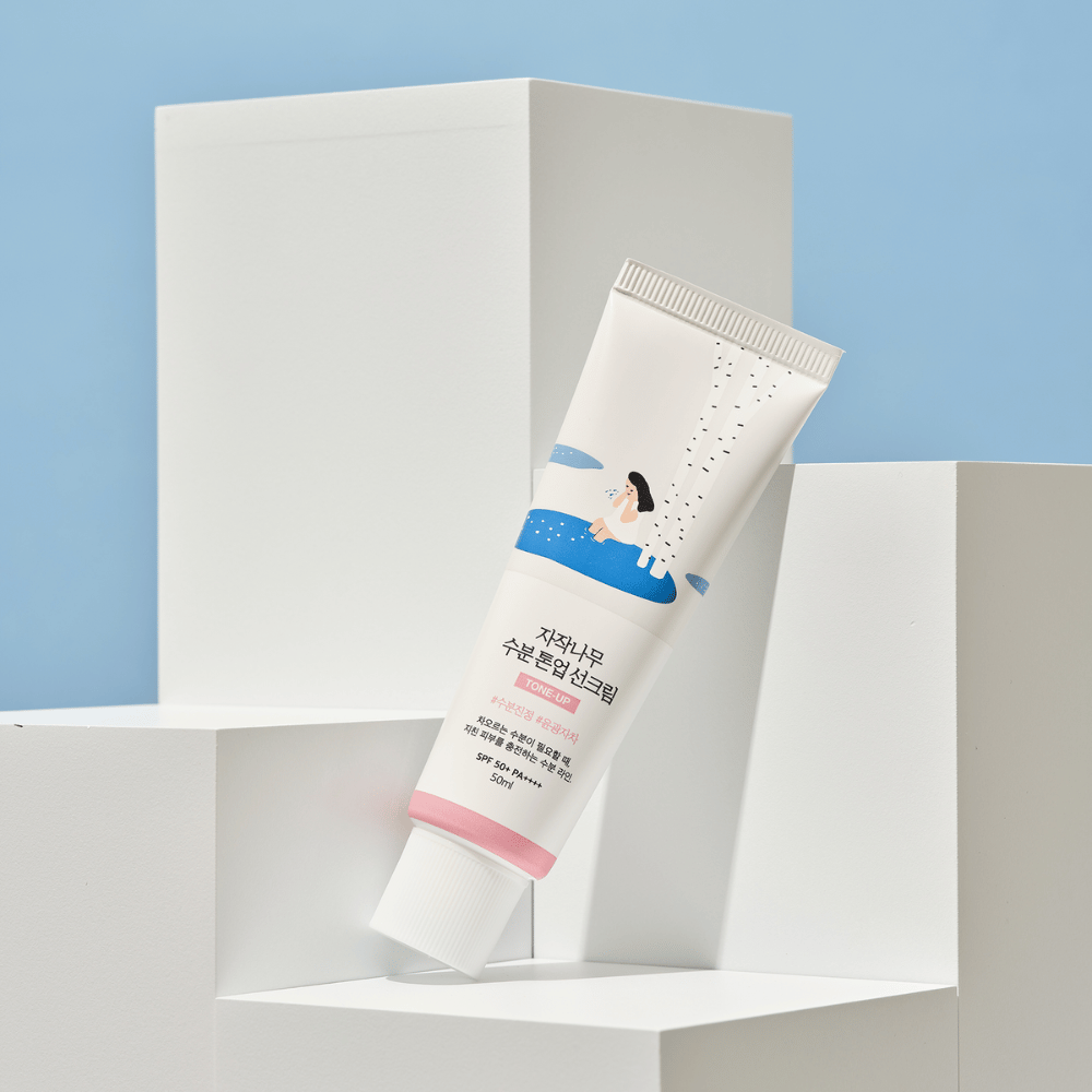 Un tubo con Birch Juice Moisturizing Tone Up Sunscreen colocado sobre cubos blancos contra un fondo celeste. El tubo es blanco con una ilustración de una mujer junto a un abedul y agua. El texto en coreano indica que el producto tiene SPF 50+ PA++++ y contiene 50 ml.
