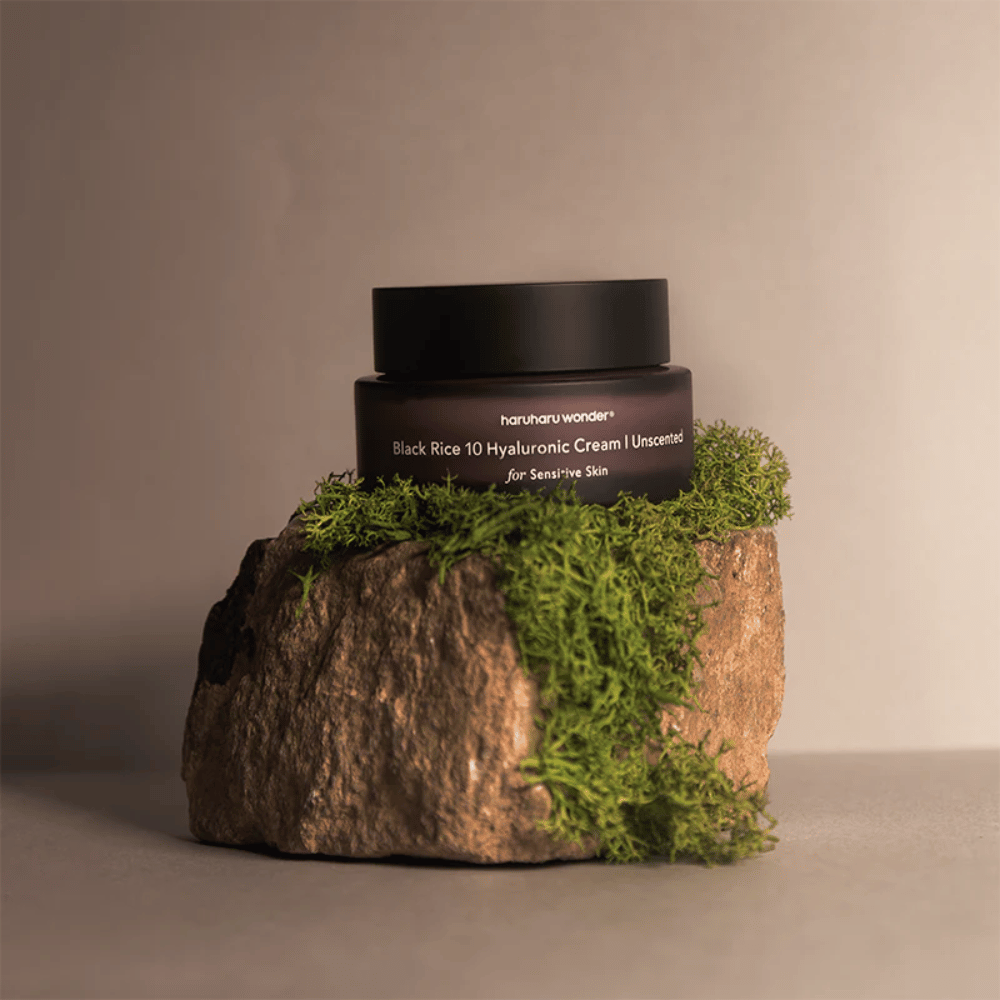 La imagen muestra un frasco de crema para la piel etiquetado "haruharu wonder Black Rice 10 Hyaluronic Cream Unscented for Sensitive Skin". El frasco es negro y está sobre una gran piedra cubierta de musgo verde claro, lo que crea una sensación natural y terrenal.