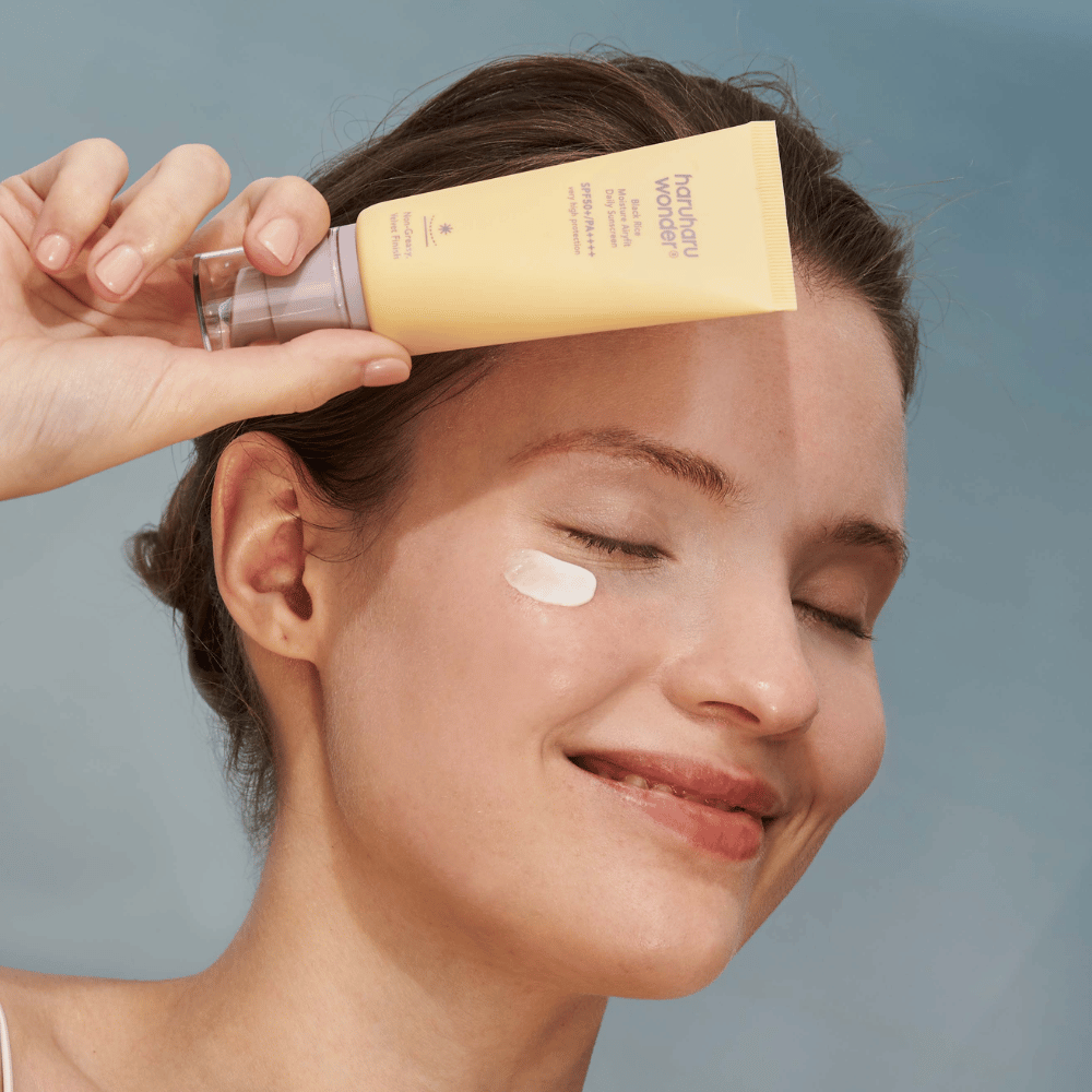 Una mujer sonriente aplica Black Rice Moisture Airyfit Daily Sunscreen SPF 50+ PA++++ de Haruharu Wonder en su rostro. Ella sostiene un tubo amarillo con protector solar y tiene un poco del producto en la mejilla. El fondo es de color azul claro.