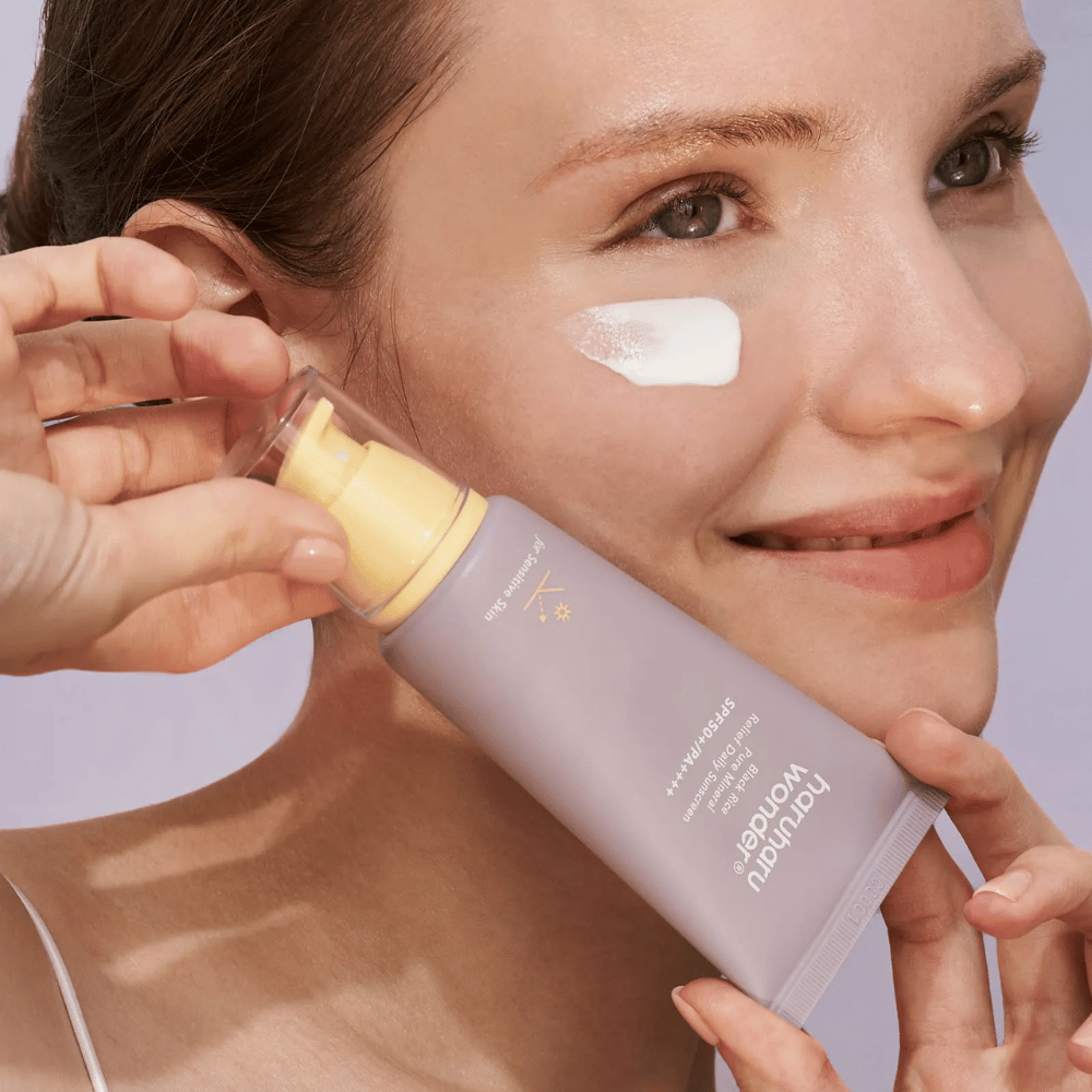Una mujer sonriente aplica Haruharu Wonder Black Rice Pure Mineral Relief Daily Sunscreen SPF 50+ PA++++ en su rostro. Ella sostiene un tubo lila con protector solar y tiene un poco del producto en la mejilla. El fondo es lila claro.