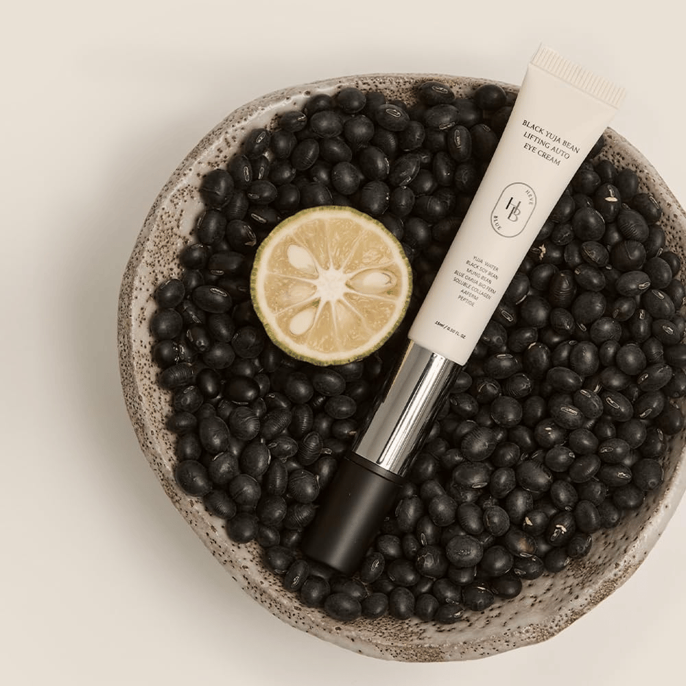 Black Yuja Bean Lifting Auto Eye Cream acostado en un tazón lleno de frijoles negros y un cítrico partido por la mitad - una representación visual de los ingredientes naturales del producto.